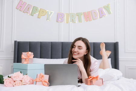 Beautiful young woman with laptop near gift boxes and rose flowers on bed in room. Happy Birthdayの写真素材