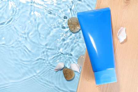 Tube of face cleansing product, stones and seashells on wooden table against light blue background,...