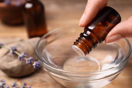 Woman dripping lavender essential oil from bottle into bowl at wooden table, closeup. Space for textの写真素材
