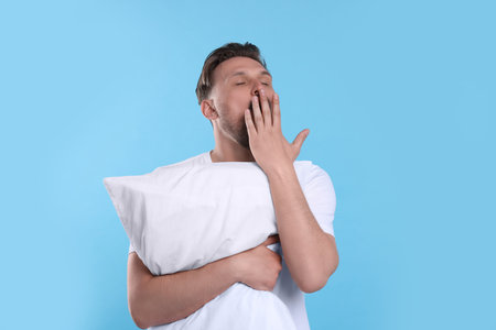 Sleepy man with pillow yawning on light blue background. Insomnia problemの写真素材