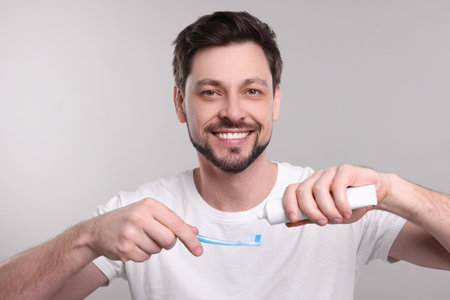 Happy man squeezing toothpaste from tube onto toothbrush on light grey backgroundの写真素材
