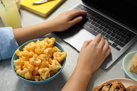 Bad eating habits. Woman using laptop surrounded by different snacks at light gray marble table, closeupの写真素材
