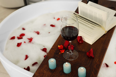 Wooden board with glass of wine, book, burning candles and rose petals on bath tubの写真素材