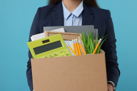 Unemployed woman holding box with personal office belongings on light blue background, closeupの写真素材