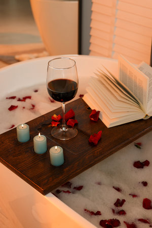 Wooden board with glass of wine, book, burning candles and rose petals on bath tubの写真素材