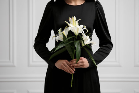 Woman with lilies near white wall, closeup. Funeral ceremonyの写真素材