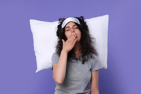 Tired young woman with sleep mask and pillow yawning on purple background. Insomnia problemの写真素材