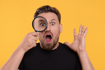 Emotional man looking through magnifier on yellow backgroundの写真素材