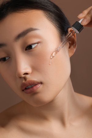 Beautiful young woman applying cosmetic serum onto her face on brown background, closeupの写真素材