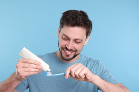 Happy man squeezing toothpaste from tube onto plastic toothbrush on light blue backgroundの写真素材