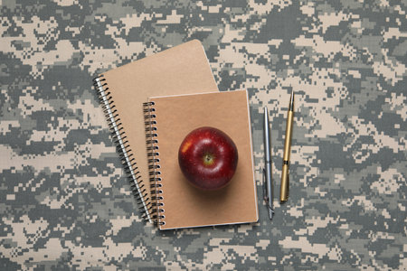 Notebooks, pens and apple on camouflage background, flat lay. Military educationの写真素材