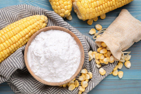 Bowl with corn starch, ripe cobs and kernels on light blue wooden table, flat lay