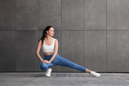 Smiling woman in sportswear stretching near dark gray wall outdoorsの写真素材
