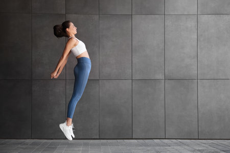 Beautiful woman in sportswear jumping near dark gray wall outdoors. Space for text