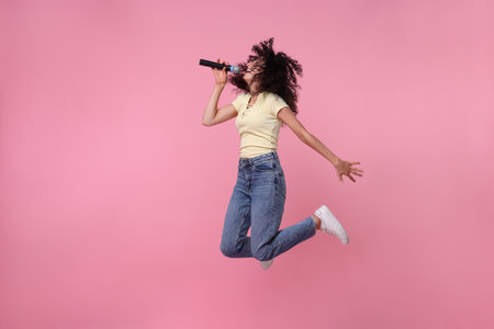 Beautiful young woman with microphone singing and jumping on pink backgroundの写真素材