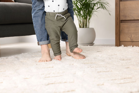 Mother supporting her baby son while he learns to walk on carpet at home, closeup