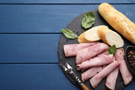 Rolled slices of delicious ham and baguette served on blue wooden table, flat lay. Space for text