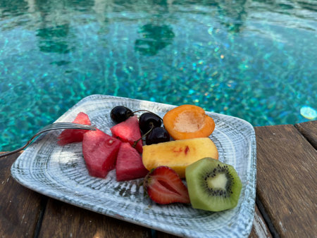 Plate with fresh fruits on wooden deck near outdoor swimming pool. Luxury resortの写真素材