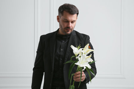 Sad man with lily flowers near white wall. Funeral ceremonyの写真素材