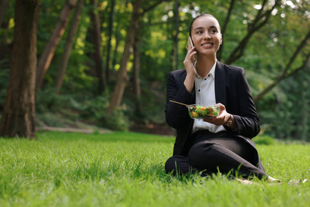 Lunch time. Happy businesswoman with container of salad talking on smartphone on green grass in park, space for textの写真素材