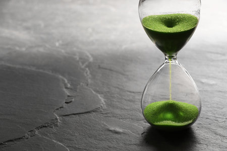 Hourglass with light green flowing sand on black table. Space for text