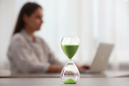Hourglass with light green flowing sand on table. Woman using laptop indoors, selective focusの写真素材
