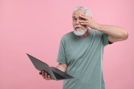 Embarrassed senior man holding laptop on pink background. Space for textの写真素材