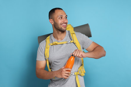 Happy tourist with backpack and thermo bottle on light blue background