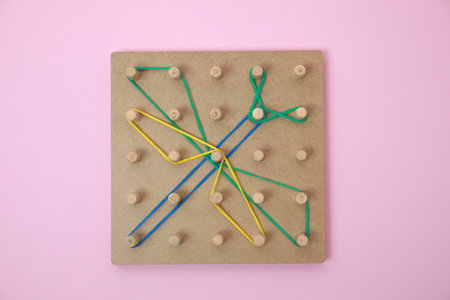 Wooden geoboard with dragonfly shape made of rubber bands on pink background, top view. Educational...