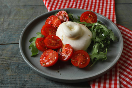 Delicious burrata cheese with tomatoes and arugula on gray wooden table, closeup