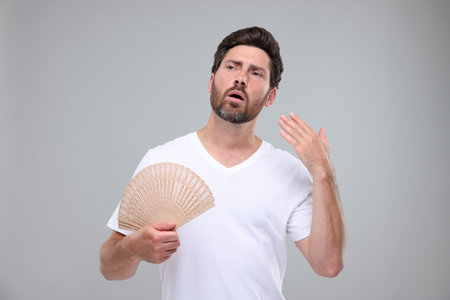 Unhappy man with hand fan suffering from heat on light gray backgroundの写真素材