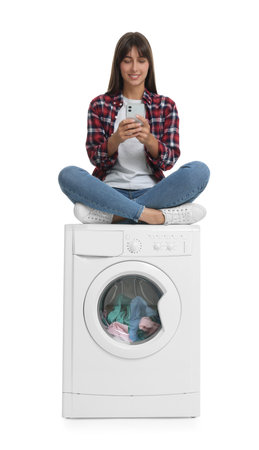Beautiful woman using smartphone while sitting on washing machine with laundry against white...