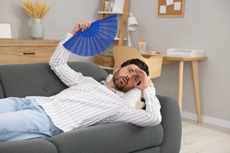 Bearded man waving blue hand fan to cool himself on sofa at homeの写真素材