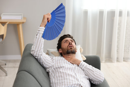 Bearded man waving blue hand fan to cool himself on sofa at homeの写真素材