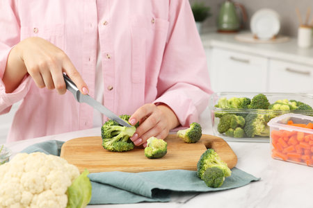 Woman cutting green broccoli near containers with fresh products at white marble table in kitchen, closeupの写真素材