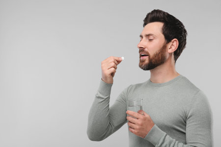 Handsome man with glass of water taking pill on light gray background, space for text