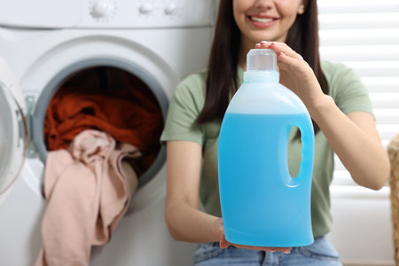 Woman holding fabric softener near washing machine with dirty clothes, closeup. Space for textの写真素材