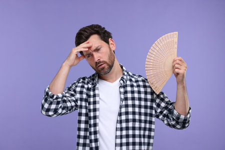 Unhappy man with hand fan suffering from heat on purple backgroundの写真素材
