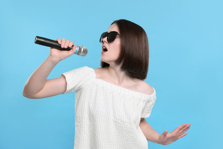 Beautiful young woman with sunglasses and microphone singing on light blue backgroundの写真素材