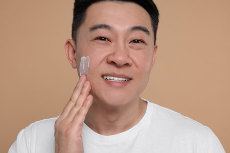 Handsome man applying cream onto his face on light brown backgroundの写真素材