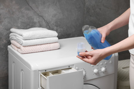 Woman pouring fabric softener from bottle into cap near washing machine indoors, closeupの写真素材