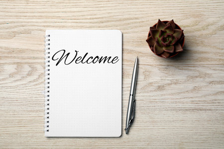 Notebook with the word Welcome, pen and houseplant on wooden table, flat lay