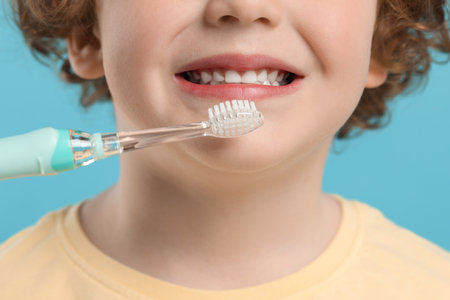Cute little boy brushing his teeth with electric toothbrush on light blue background, closeup