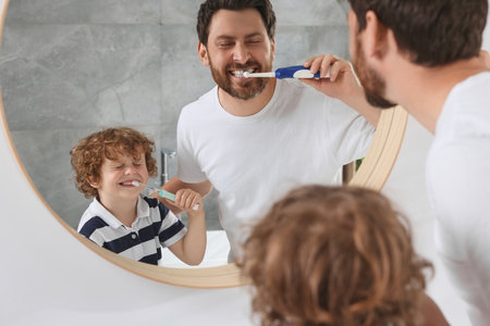 Father and his son brushing teeth together near mirror indoors