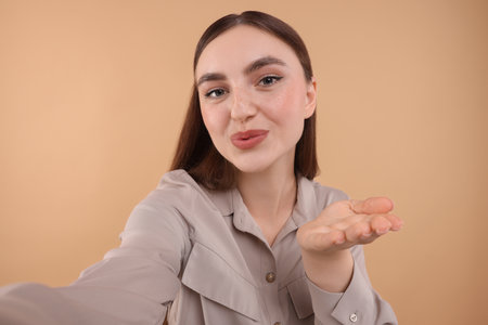 Beautiful woman taking selfie and blowing kiss on beige backgroundの写真素材