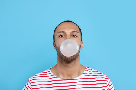 Portrait of young man blowing bubble gum on light blue backgroundの写真素材