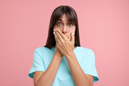 Embarrassed woman covering mouth with hands on pink backgroundの写真素材