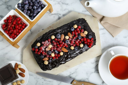 Delicious chocolate sponge cake, tea and ingredients on white marble table, flat lay