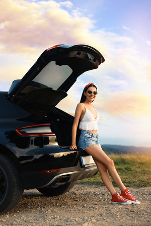 Happy woman sitting in trunk of modern car on roadside outdoorsの写真素材