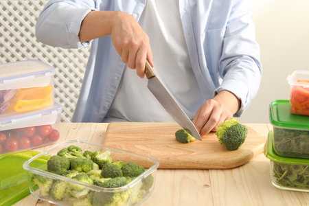 Man cutting fresh broccoli with knife near containers at wooden table, closeup. Food storageの写真素材